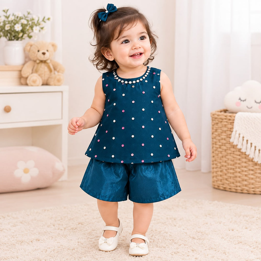 A toddler girl wearing a dark teal sleeveless top with white, pink, and blue polka dots and matching teal shorts, standing in a bright, neutral nursery setting