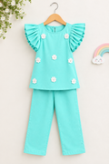 A toddler girl's two-piece outfit set in mint green, featuring a short-sleeve top with ruffled sleeves and white fabric flowers, paired with matching pants, hanging on a wooden hanger against a light beige background.