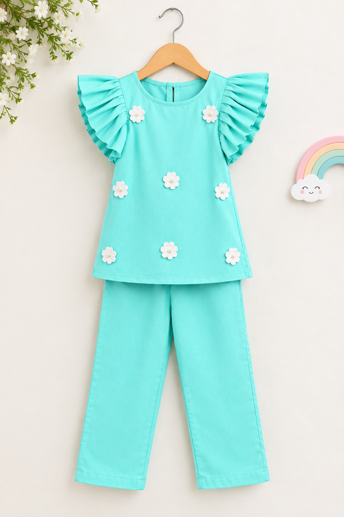A toddler girl's two-piece outfit set in mint green, featuring a short-sleeve top with ruffled sleeves and white fabric flowers, paired with matching pants, hanging on a wooden hanger against a light beige background.