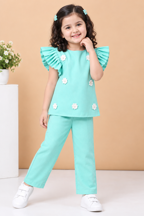A toddler girl's two-piece outfit set in mint green, featuring a short-sleeve top with ruffled sleeves and white fabric flowers, paired with matching pants, hanging on a wooden hanger against a light beige background.